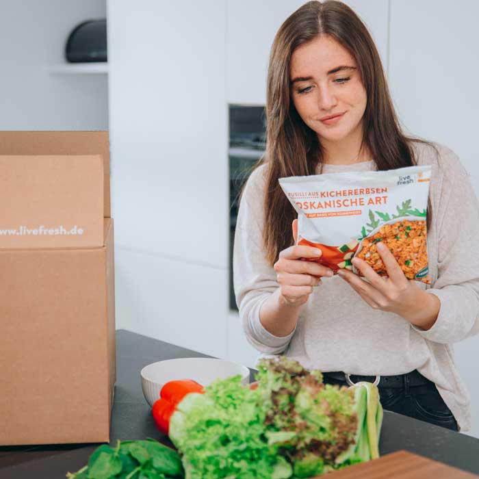 Eine Frau in einer modernen Küche, die eine Verpackung mit der Aufschrift „Kichererbsen Toskansiche Art“ in der Hand hält und interessiert betrachtet. Auf der Arbeitsfläche vor ihr liegen frische Zutaten wie Salat, Paprika und Sellerie. Neben ihr steht ein Paket mit der Aufschrift „livefresh.de“.