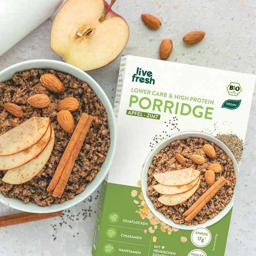 Zeigt eine Packung „Live Fresh“ Porridge in der Geschmacksrichtung Apfel-Zimt, daneben eine Schale mit dem zubereiteten Porridge, garniert mit Apfelscheiben, Mandeln und einer Zimtstange. Die Verpackung weist darauf hin, dass das Produkt vegan, proteinreich und bio-zertifiziert ist, mit Zutaten wie Sojaflocken, Chiasamen und Hanfsamen. Eine aufgeschnittene Apfelhälfte und verstreute Mandeln ergänzen die Szene.