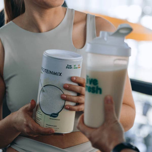 Zeigt eine Person in Sportkleidung, die eine Dose „Live Fresh Proteinmix“ in der Hand hält. Eine zweite Person reicht ihr einen Shake in einem „Live Fresh“-Shaker. Das Bild vermittelt eine aktive und gesundheitsbewusste Szene, die den Proteinshake als Teil einer Fitness- oder Ernährungsroutine darstellt. Der Proteinmix ist vegan und als Trinkmahlzeit geeignet.