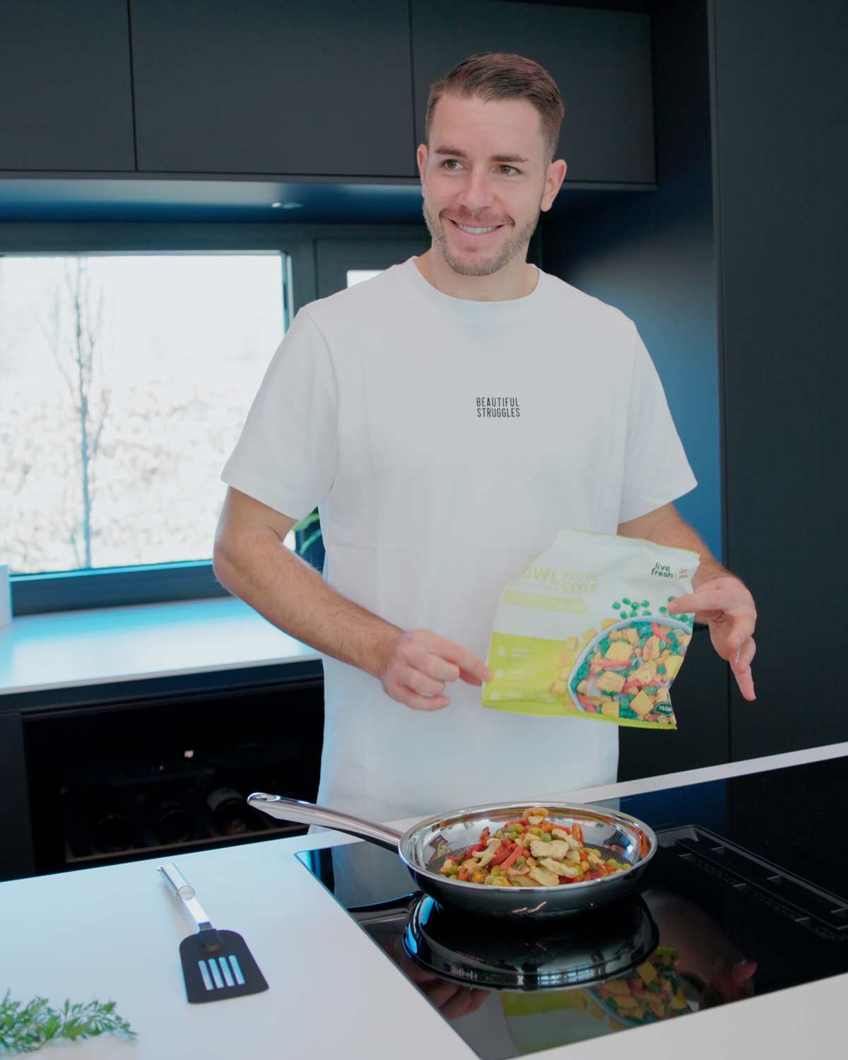 Ein Mann in einem weißen T-Shirt mit der Aufschrift "Beautiful Struggles", der in einer modernen Küche steht. Er bereitet eine Mahlzeit in einer Pfanne zu und hält eine Packung "Bowl Exotic Style" von Live Fresh in der Hand, die vegane Tiefkühlmahlzeiten enthält.