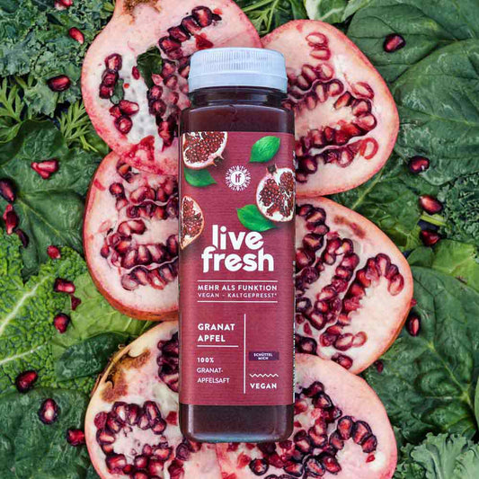 Eine Flasche „Live Fresh Granatapfel“ Saft, die auf einem Hintergrund aus halbierten Granatäpfeln und grünem Blattgemüse liegt. Der Saft ist vegan, kaltgepresst und besteht zu 100% aus Granatapfelsaft. Die Verpackung betont die Frische und Natürlichkeit des Produkts und hebt hervor, dass es ohne Zusatzstoffe und in einer schonenden Verarbeitung hergestellt wird.