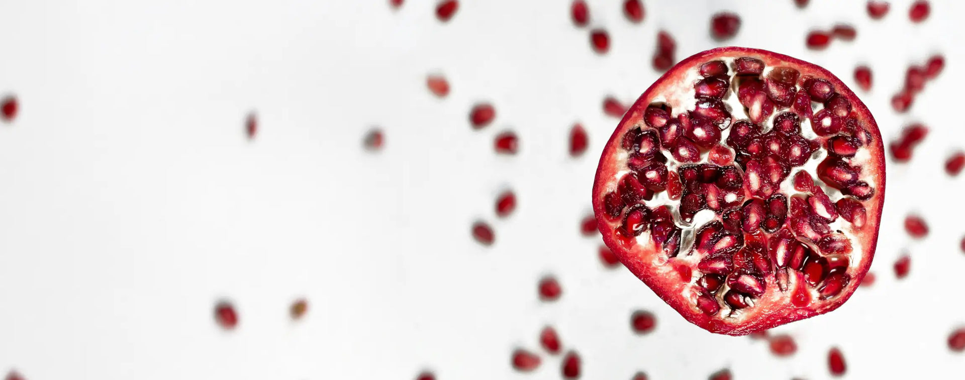Halber Granatapfel auf weißem Hintergrund mit sichtbar leuchtend roten Kernen. Einzelne Kerne liegen locker um die Frucht verteilt.