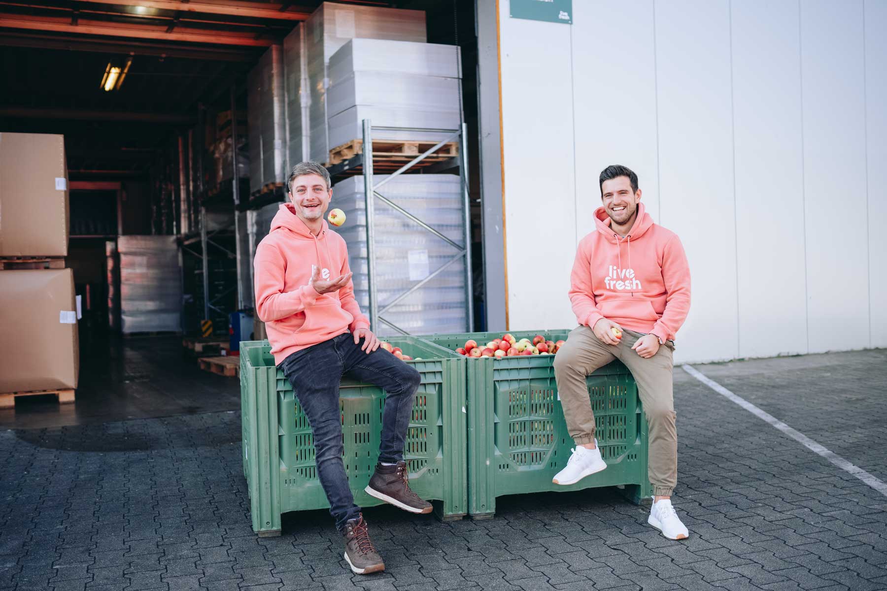 Zeigt zwei Männer in rosafarbenen Hoodies mit der Aufschrift „Live Fresh“, die auf großen grünen Kisten voller Äpfel sitzen. Beide lächeln, während einer der Männer einen Apfel in der Hand hält und der andere einen Apfel in die Luft wirft. Sie befinden sich vor einer Lagerhalle oder Produktionsstätte, was auf die Verbindung zu frischen Zutaten und die Herstellung von Live Fresh-Produkten hinweist. Die Stimmung ist fröhlich und entspannt, was auf die Leidenschaft für ihr Produkt und ihre Marke hindeutet.