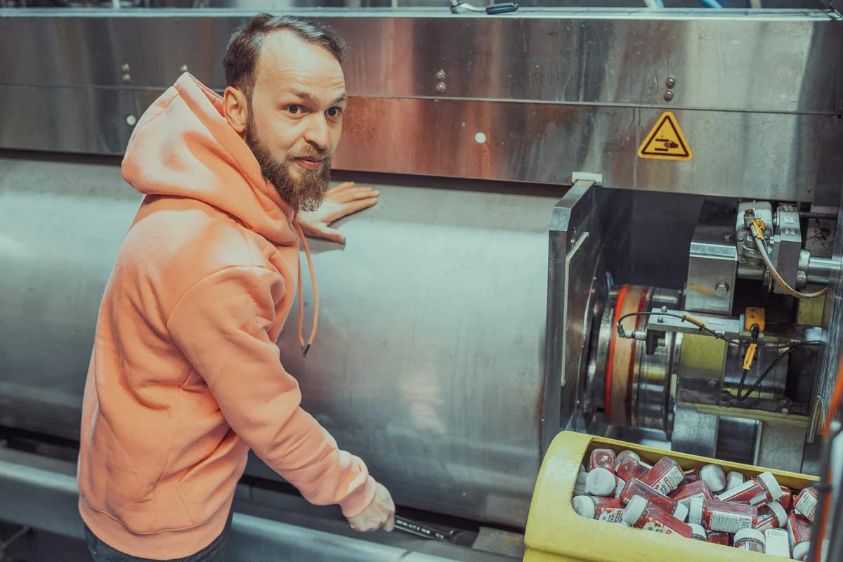 Mann in einem orangefarbenen "Live Fresh"-Hoodie, der neben einer industriellen Maschine arbeitet. Im Vordergrund ist eine gelbe Vorrichtung mit mehreren kleinen Flaschen gefüllt. Der Mann scheint die Maschine zu überwachen oder anzupassen. Die Szene vermittelt den Prozess der Herstellung oder Verarbeitung von Produkten, vermutlich Säfte oder Shots, in einem industriellen Umfeld.