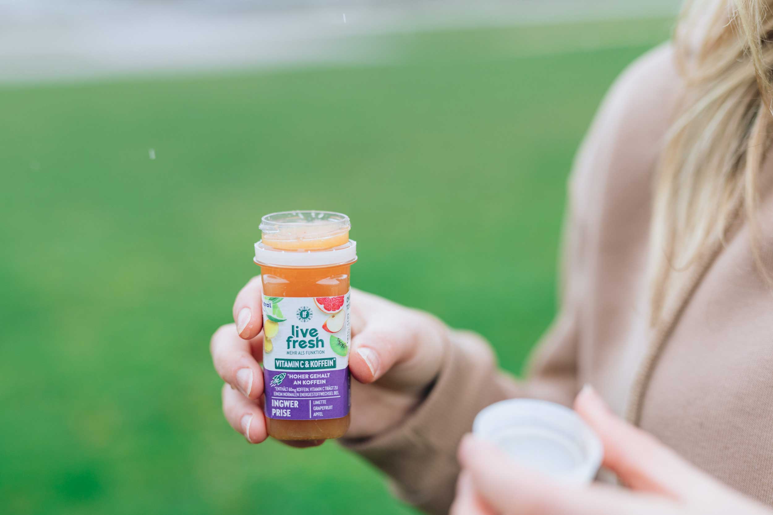 Person hält eine geöffneten Shot namens Ingwer Prise von LiveFresh in der Hand, vor grünem Hintergrund im Freien.