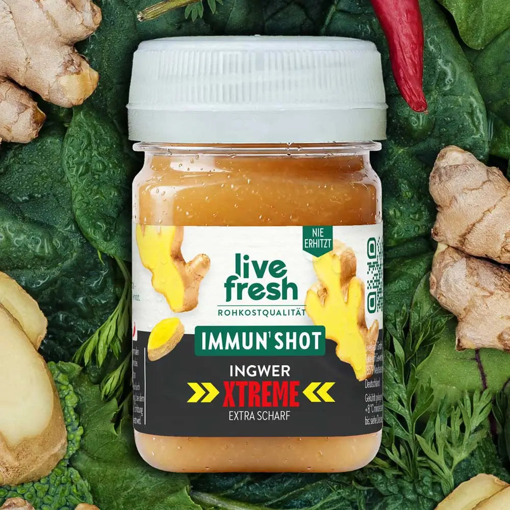 Ein Live Fresh Immun Shot der Sorte Ingwer Xtreme steht zwischen frischen Ingwerwurzeln, Ingwerscheiben und roten Chilischoten auf dunkelgrünem Blattgemüse. Auf dem Etikett stehen die Aufschriften "IMMUN SHOT", "INGWER XTREME" und "EXTRA SCHARF".