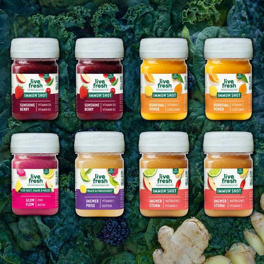 Acht bunte LiveFresh Shots in zwei Reihen auf dunklem Blattgemüse mit Ingwer und Brombeere. Die Sorten sind "Sunshine Berry Vitamin D3 Vitamin K2" "Kurkuma Power Vitamin C Curcumin" "Glow Flow Zink Biotin" "Ingwer Prise Vitamin C Koffein" und "Ingwer Sturm Natürliches Vitamin C".