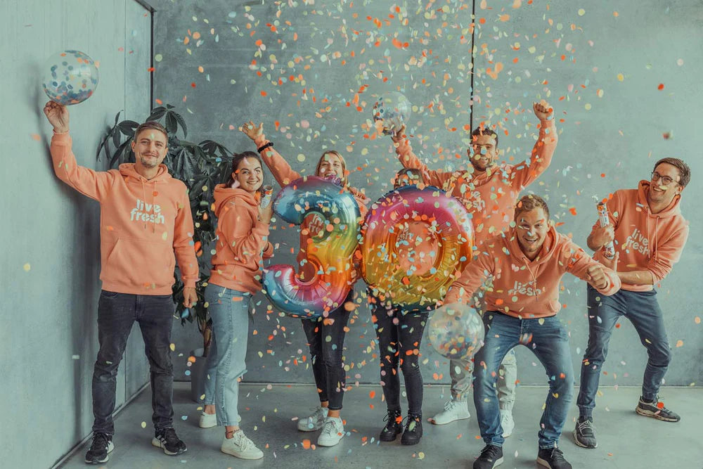 Ein fröhliches Team, das den Meilenstein von 30k feiert. Die Personen tragen "Live Fresh"-Pullover und stehen um große, bunte Ballons in der Form der Zahl "30", während Konfetti in die Luft geworfen wird. Die Stimmung ist festlich, und die Gruppe strahlt Freude und Begeisterung aus.