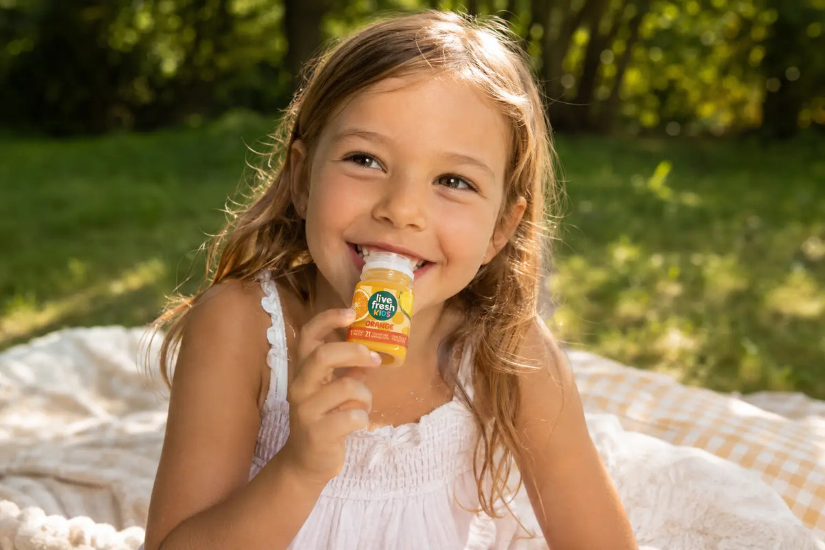 Een meisje zit glimlachend op een picknickdeken in het groen en drinkt een LiveFresh Kids-sinaasappelsap. 