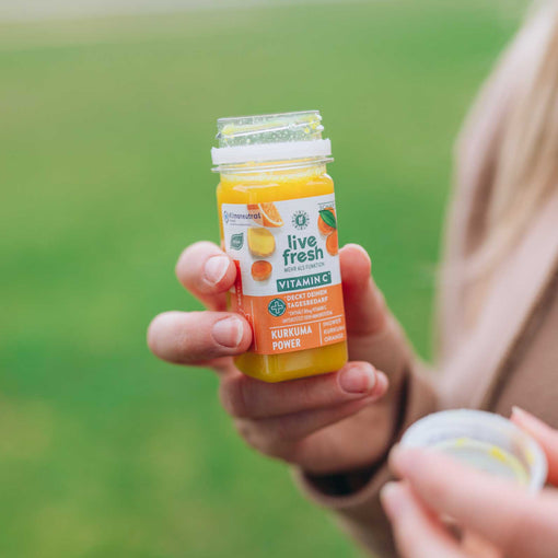 Eine Person, die einen Kurkuma Power Shot von Live Fresh in der Hand hält. Der Shot verspricht Vitamin C und enthält Zutaten wie Ingwer, Kurkuma und Orange. Die Person hält den geöffneten Deckel in der anderen Hand, während sie den Shot draußen, vor einem grünen Hintergrund, genießt.