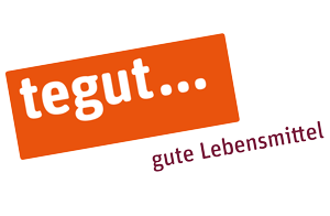 Das Logo von Tegut, einem deutschen Lebensmittelhändler. Das Logo besteht aus einem orangefarbenen Rechteck mit dem weißen Schriftzug „tegut…“, darunter steht in kleineren, burgunderroten Buchstaben „gute Lebensmittel“.