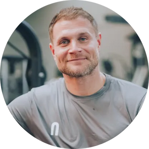 Mimi Kraus; lächelnd, in einem grauen T-Shirt sportlich gekleidet mit kurzem Bart und hellbraunen Haaren in einem Fitnessstudio, im Hintergrund sind Trainingsgeräte unscharf sichtbar.