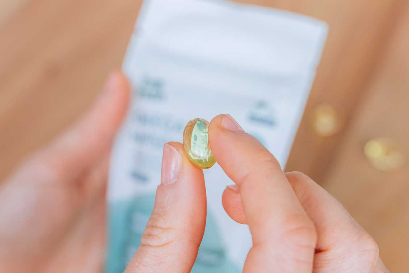 Nahaufnahme von zwei Händen, die eine durchsichtige, gelbliche Omega-3-Kapsel halten. Im Hintergrund ist unscharf eine Verpackung zu erkennen, die möglicherweise weitere Informationen zu dem Produkt enthält. Die Aufnahme betont die Beschaffenheit und Details der Kapsel, die leicht glänzt.