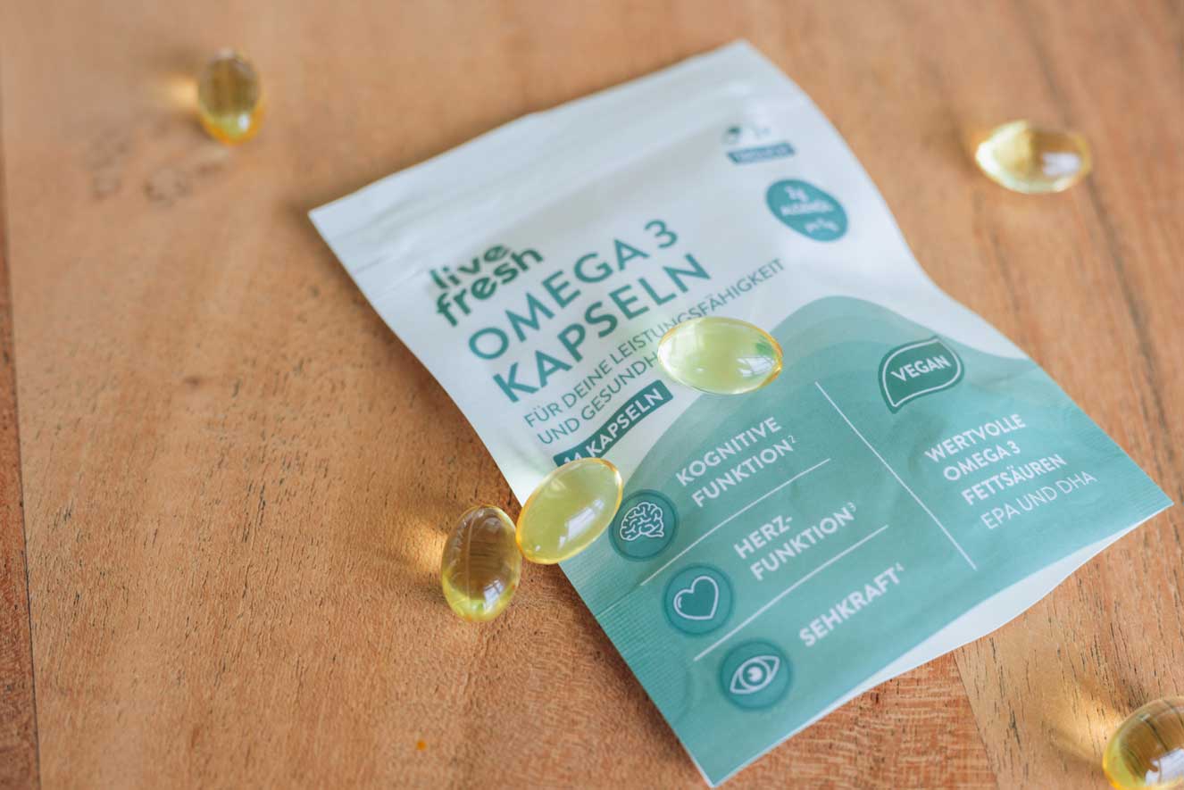 Verpackung von „Live Fresh Omega 3 Kapseln“ auf einer Holzoberfläche. Rund um die Verpackung liegen mehrere durchsichtige, gelbliche Omega-3-Kapseln verstreut. Auf der Verpackung sind Informationen über die Vorteile der Kapseln aufgeführt, darunter „Kognitive Funktion“, „Herzfunktion“ und „Sehkraft“. Zudem wird hervorgehoben, dass das Produkt vegan ist und wertvolle Omega-3-Fettsäuren, einschließlich EPA und DHA, enthält.