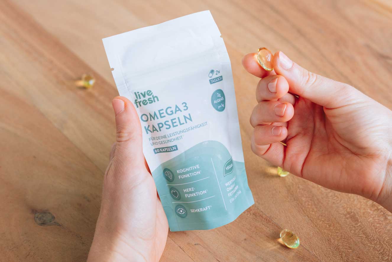 Zwei Hände, die eine Verpackung von „Live Fresh Omega 3 Kapseln“ halten. Eine der Hände hält zudem eine einzelne, durchsichtige Omega-3-Kapsel zwischen den Fingern. Auf der Verpackung sind wichtige Informationen zum Produkt zu sehen, wie die Förderung der „Kognitiven Funktion“, „Herzfunktion“ und „Sehkraft“. Zudem wird darauf hingewiesen, dass die Kapseln vegan sind und wertvolle Omega-3-Fettsäuren enthalten.