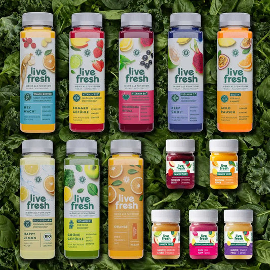 Probierpaket alle Säfte und Shots von LiveFresh mit verschiedenfarbigen Säften und kleinen Immun Shots. Die Flaschen sind auf frischem grünem Blattgemüse angeordnet und zeigen Sorten wie Happy Lemon. Grüne Gefühle. Orange. Sommer Gefühle. Keep Cool und Gold Rausch.