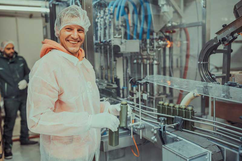 Ein Mitarbeiter in Schutzkleidung und mit Haarnetz steht lächelnd an einer Produktionslinie und hält eine Flasche Live Fresh Selleriesaft. Im Hintergrund sieht man weitere Flaschen, die abgefüllt werden. Das Bild wirkt professionell und vermittelt Vertrauen in die hygienische und saubere Produktionsweise des Produkts. 