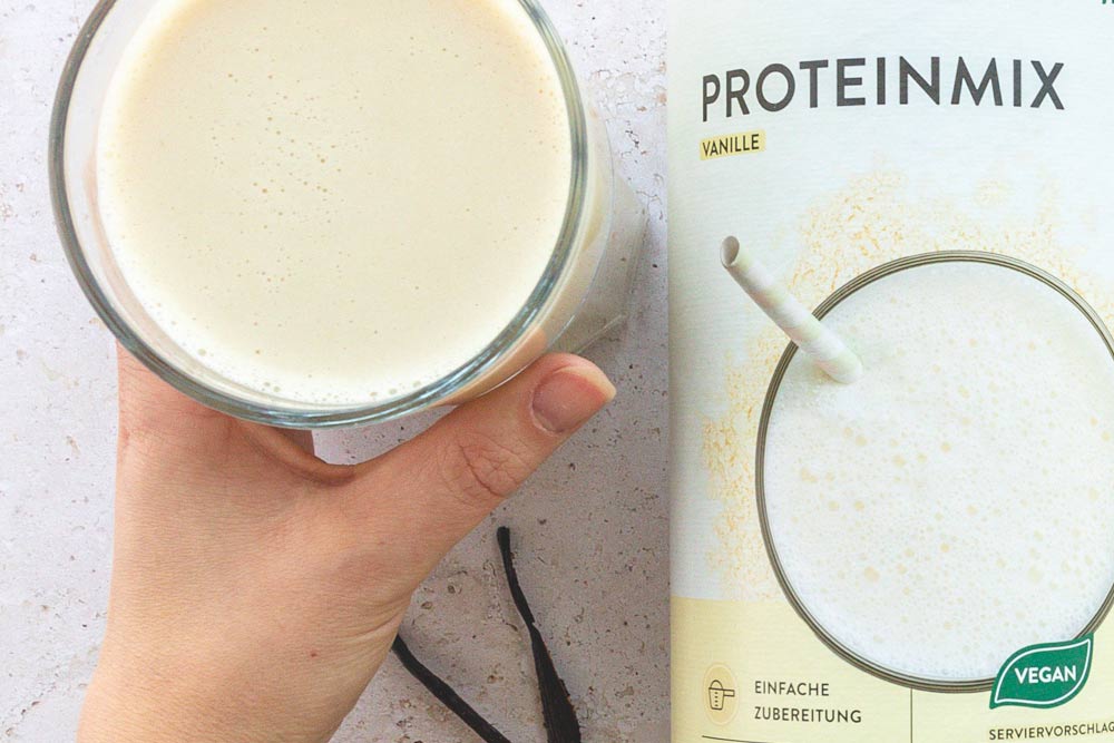 Eine Hand, die ein Glas mit einem cremigen Proteinshake hält, daneben ist eine Packung des veganen Proteinmixes in der Geschmacksrichtung Vanille zu sehen. Der Shake sieht glatt und perfekt gemixt aus, und die Verpackung unterstreicht die einfache Zubereitung sowie die vegane Natur des Produkts. Das Bild wirkt frisch und ansprechend, es lädt dazu ein, den Shake als Teil einer gesunden Ernährung zu genießen.