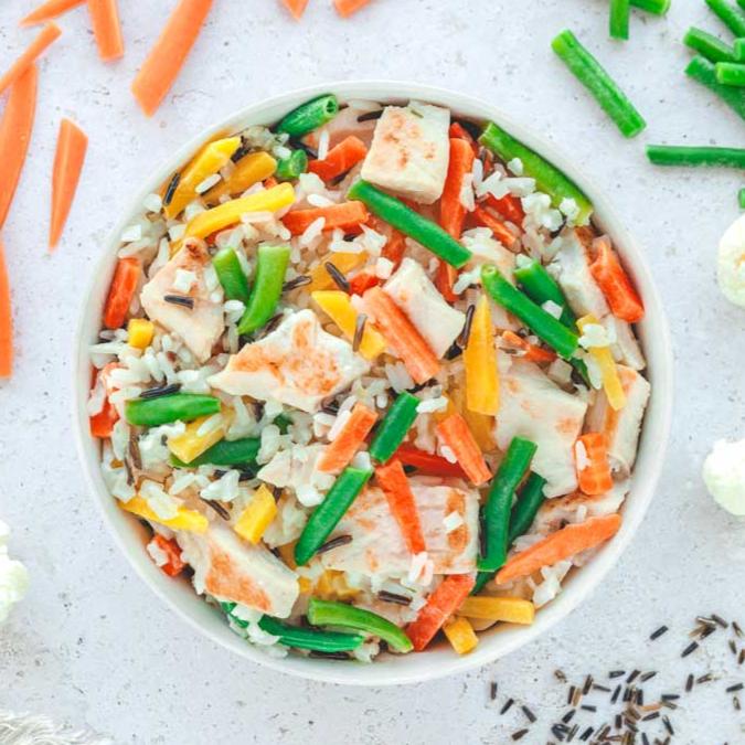 Ein weiteres Bild zeigt die angerichtete Reis-Bowl mit Hähnchenstücken, Karottenstreifen, grünen Bohnen und buntem Gemüse auf Wildreis. Die um den Teller verstreuten frischen Zutaten, darunter Karotten und Blumenkohl, verleihen dem Gericht ein frisches und farbenfrohes Aussehen.