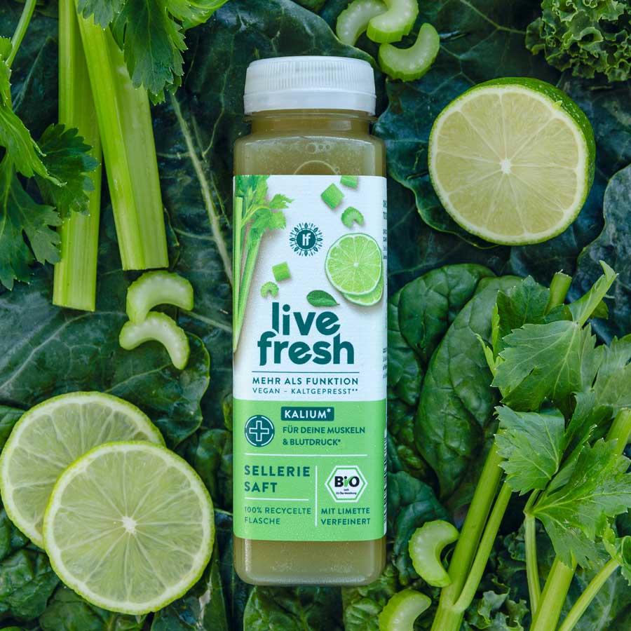 Das Bild zeigt eine Flasche "Live Fresh" Selleriesaft, die auf einem Hintergrund aus frischen Selleriestangen, Limettenscheiben und Grünkohlblättern liegt. Der Saft ist vegan, kaltgepresst und bio-zertifiziert. Auf dem Etikett steht „Mehr als Funktion“ und „Kalium für deine Muskeln & Blutdruck“. Der Saft enthält zusätzlich Limette und wird in einer 100 % recycelten Flasche angeboten.