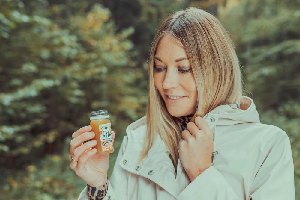 unge Frau mit blonder Frisur und heller Jacke steht im Grünen und hält einen kleinen LiveFresh-Shot in der Hand, während sie lächelnd darauf blickt.
