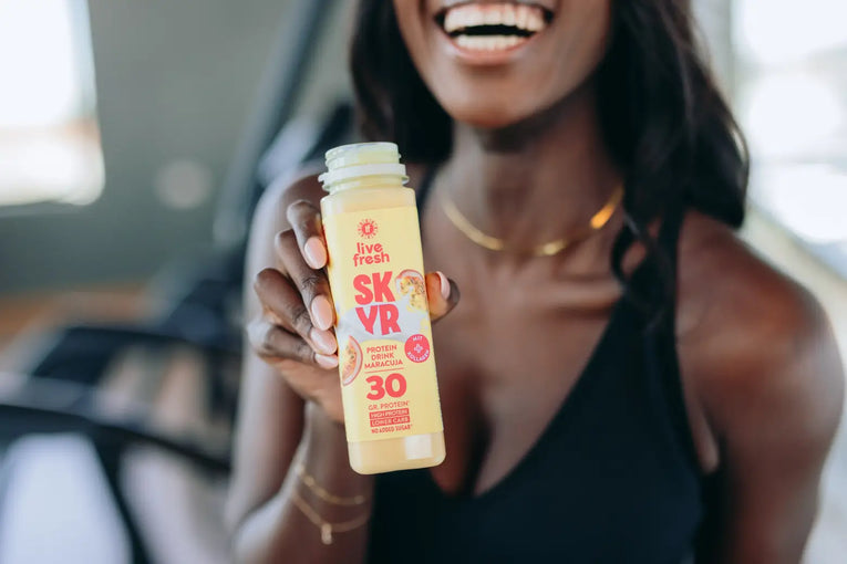 Frau im Fitnessstudio hält eine geöffnete Flasche LiveFresh SKYR Protein Drink Maracuja mit 30 Gramm Protein in die Kamera.
