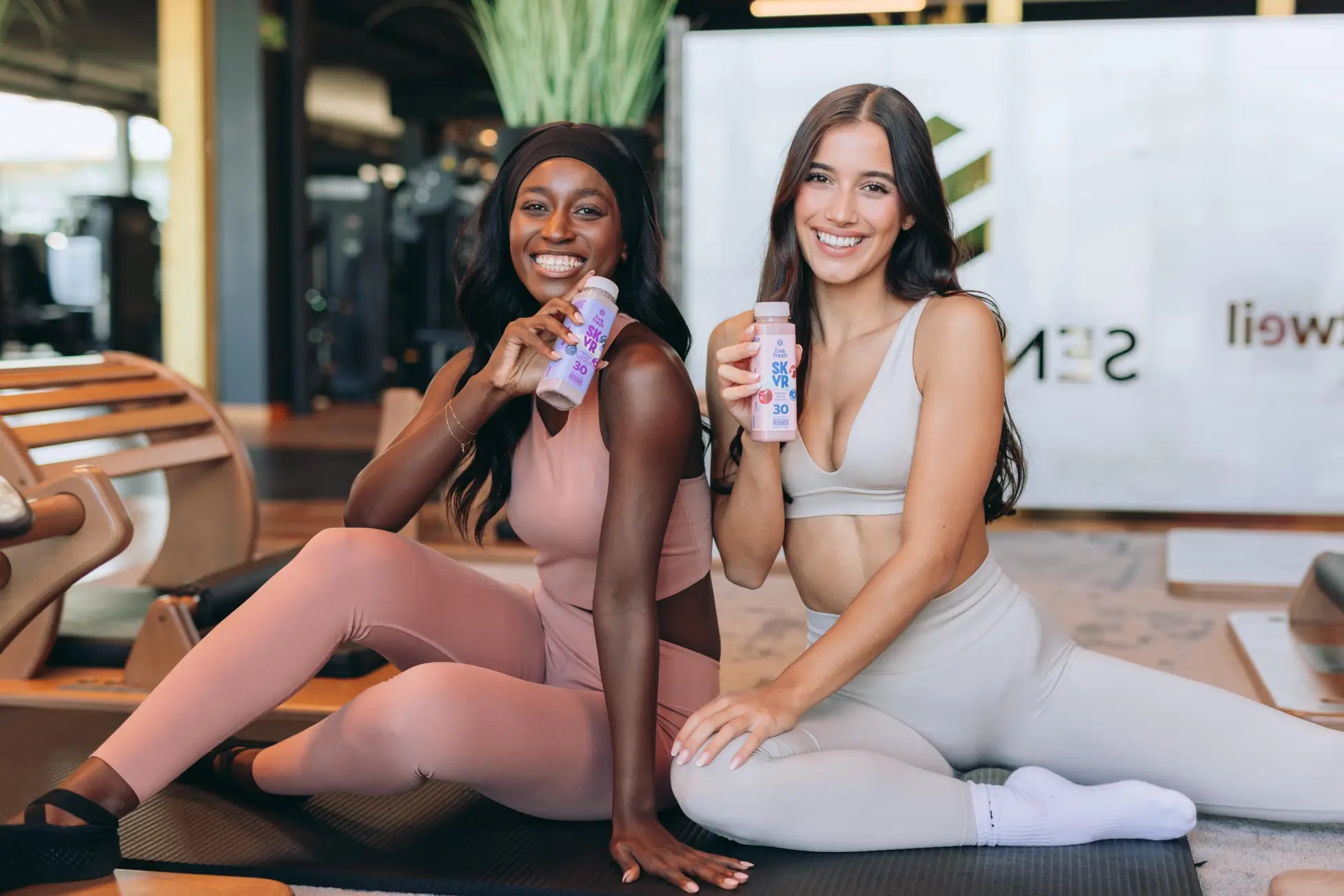 Zwei Frauen in Sportkleidung sitzen auf Trainingsmatten im Fitnessstudio und halten lächelnd ihre Proteinshake Flaschen in die Kamera.