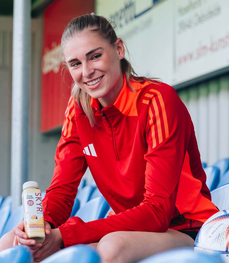 Eine sportliche Frau im roten Trainingsanzug sitzt auf einer Tribüne und hält einen Skyr Protein Drink mit Maracuja-Geschmack von Live Fresh, der 30g Protein enthält.