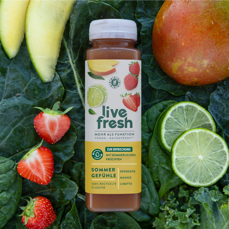 eine Flasche "Live Fresh Sommer Gefühle" Saft, umgeben von frischen Erdbeeren, Mangoscheiben, Limettenscheiben und Grünkohlblättern. Der Saft hebt die sommerliche Erfrischung durch die Zutaten Erdbeere, Mango und Limette hervor. Die Flasche besteht aus 100 % recyceltem Material und ist vegan sowie kaltgepresst.