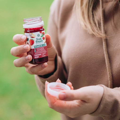 Person hält einen geöffneten Vitamin-D3+K2 Shot namens Sunshine Berry der Marke LiveFresh in der Hand, im Hintergrund ist ein Feld zu erkennen. 