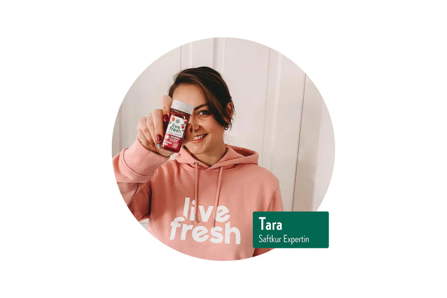 Eine Frau, die einen „Live Fresh“-Shot in die Kamera hält. Sie trägt einen rosa Hoodie mit dem „Live Fresh“-Logo. Ihr Lächeln und die Geste, das Produkt zu präsentieren, vermitteln Selbstbewusstsein und Zufriedenheit. Die Darstellung wirkt freundlich und einladend, passend zum Thema gesunde Ernährung oder Saftkuren. Der grüne Balken mit dem Text „Tara – Saftkur Expertin“ betont ihre Rolle als Botschafterin oder Expertin für das Produkt.