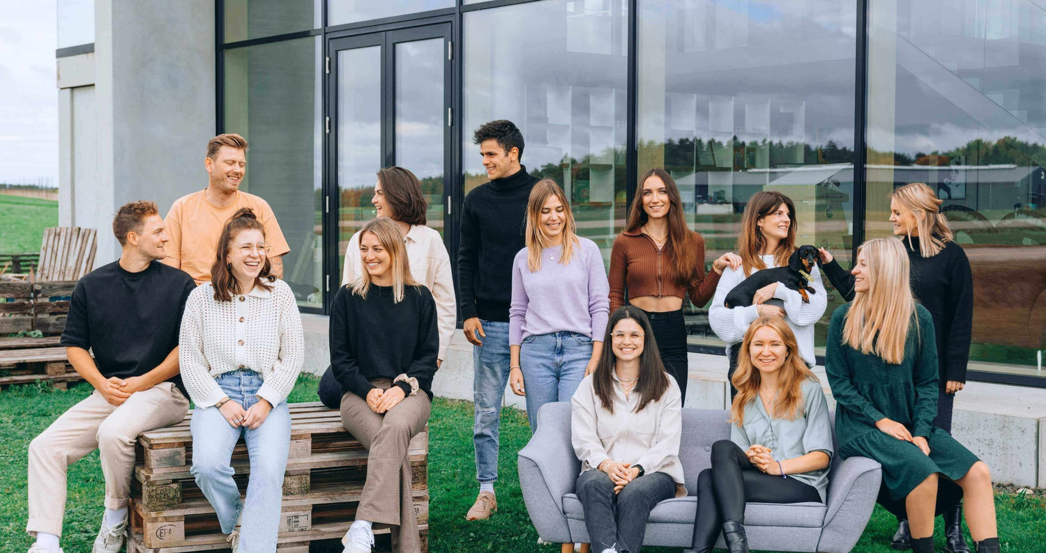 Eine Gruppe von zwölf jungen Menschen sitzt und steht entspannt vor einem modernen Bürogebäude mit großen Glasfenstern. Sie lachen und unterhalten sich, während eine Person einen kleinen schwarzen Hund hält, der von einer anderen gestreichelt wird. Die Gruppe ist leger gekleidet, einige sitzen auf Paletten, andere auf einem grauen Sofa, mit einer grünen Wiese im Vordergrund und einem ländlichen Hintergrund.