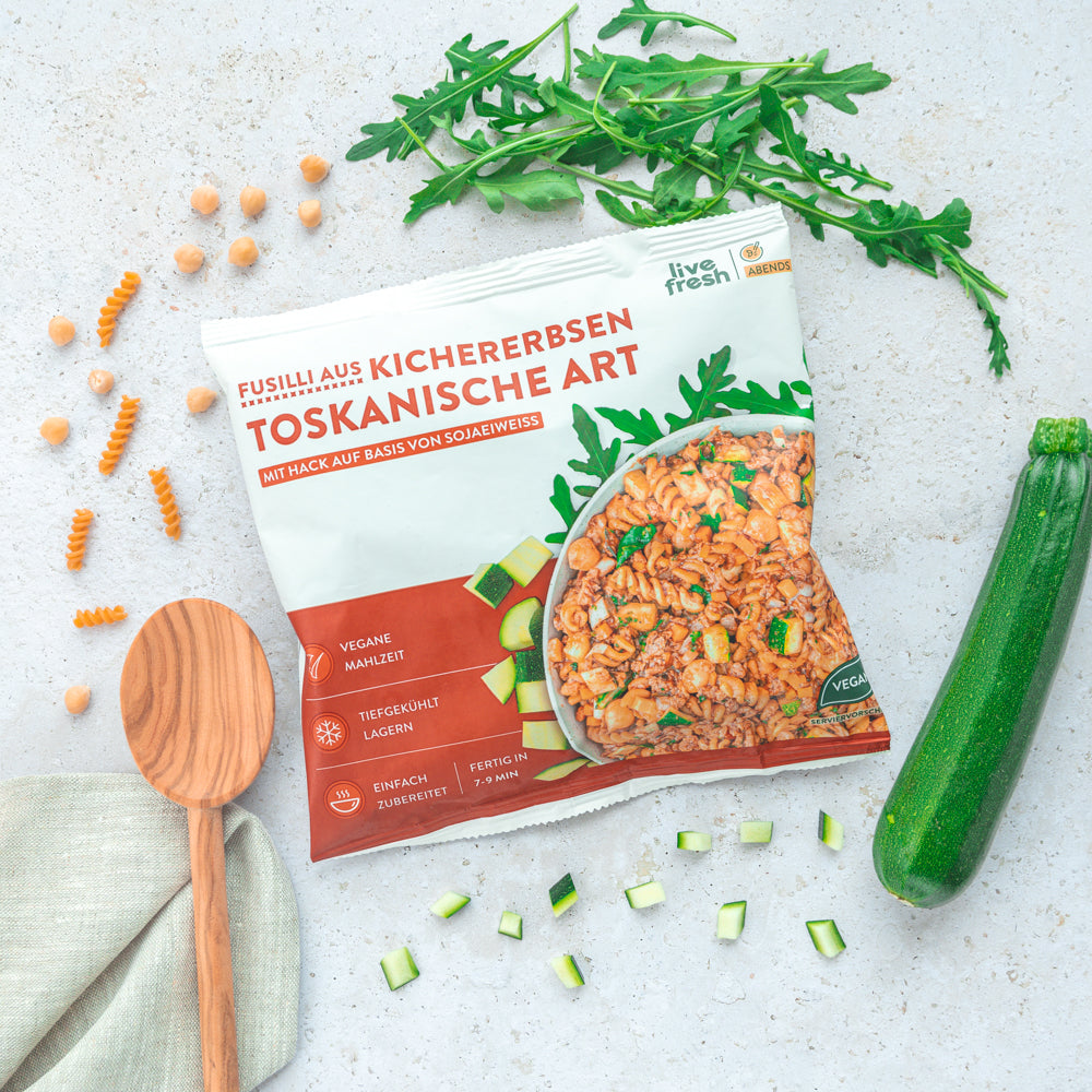 Eine Verpackung von "Fusilli aus Kichererbsen Toskanische Art" von Live Fresh, einer veganen Mahlzeit mit Sojaeiweiß als Basis. Die Verpackung ist von frischen Zutaten wie Zucchini und Rucola umgeben, was die natürliche und gesunde Komposition des Gerichts unterstreicht.