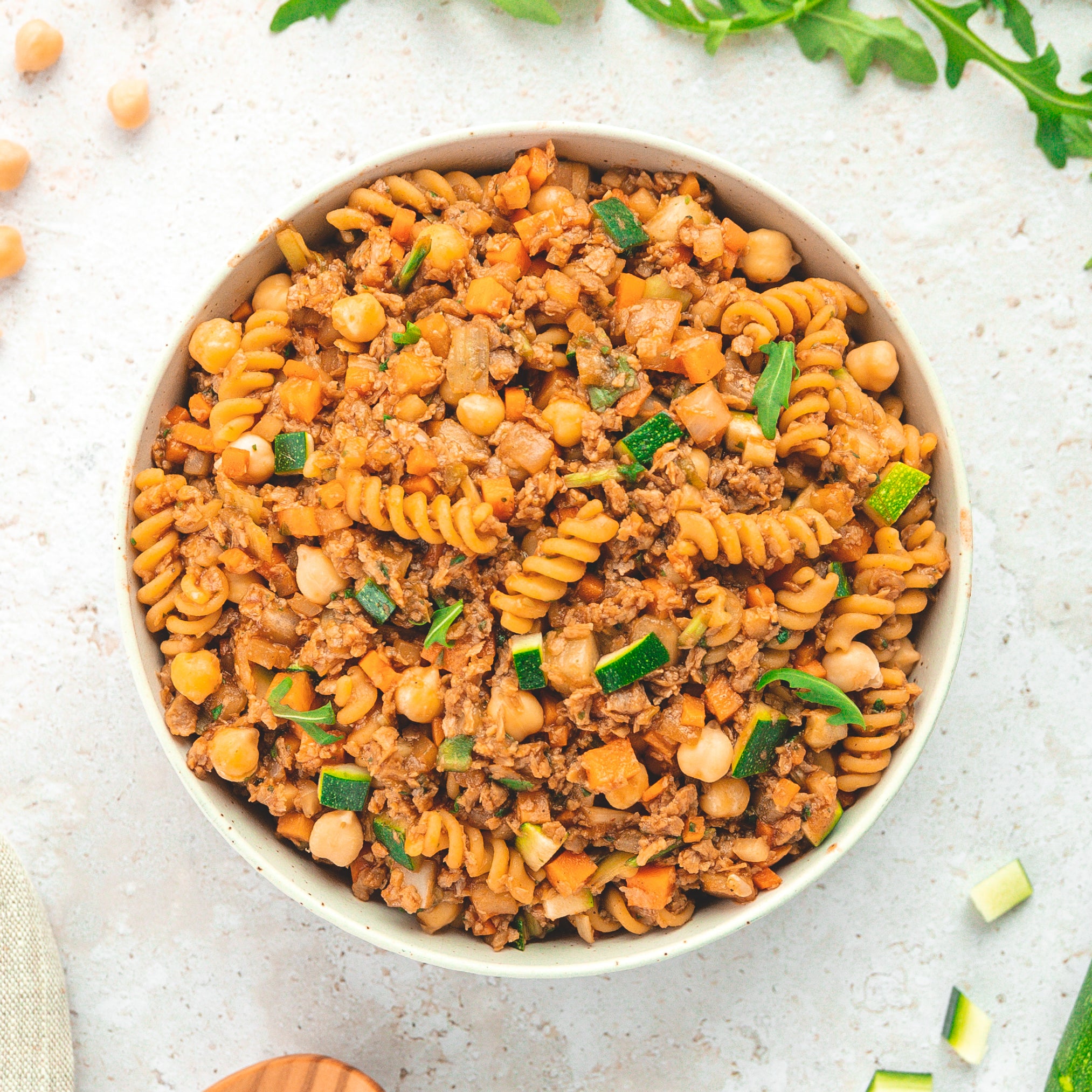 Eine Schüssel mit veganen Fusilli-Nudeln, gemischt mit einer herzhaften Sauce aus gehacktem Gemüse wie Zucchini, Karotten und Kichererbsen. Die Zutaten scheinen in einer Tomatensauce oder einer ähnlichen würzigen Mischung gekocht zu sein.