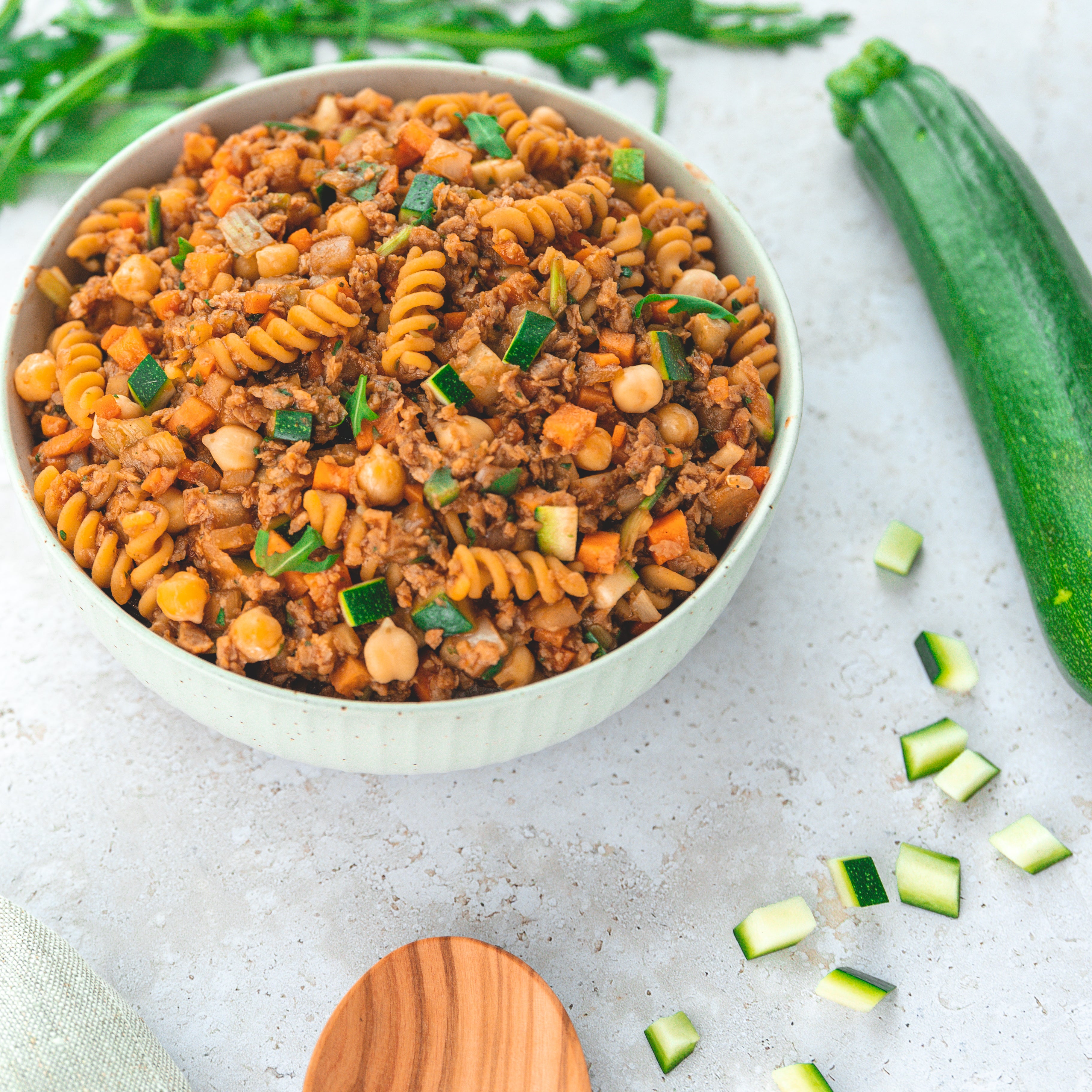 Eine Schüssel mit veganen Fusilli-Nudeln, gemischt mit Zucchiniwürfeln, Kichererbsen und anderen Gemüsesorten in einer herzhaften, pflanzlichen Sauce. Neben der Schüssel liegt eine Zucchini, die die frischen, natürlichen Zutaten betont.