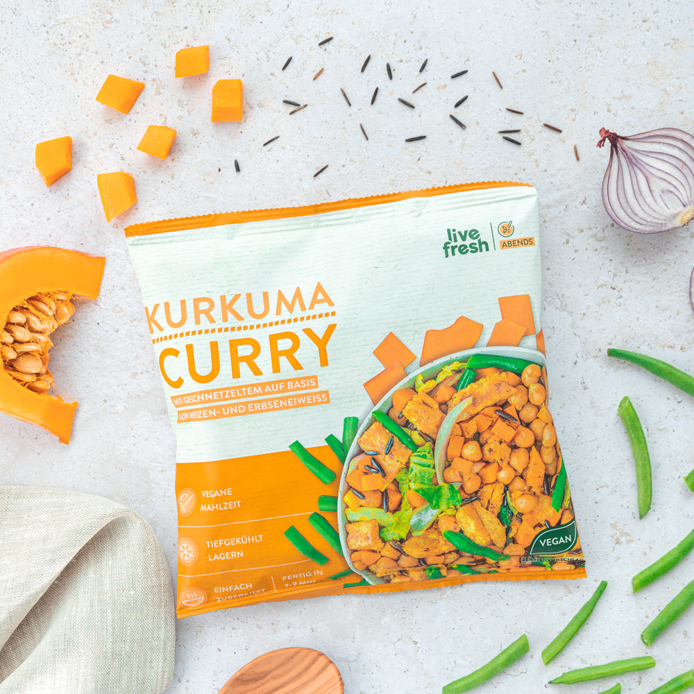 Eine Verpackung des „Kurkuma Curry“ von Live Fresh, umgeben von Kürbiswürfeln, grünen Bohnen, Wildreis und einer Zwiebelhälfte. Das vegane Gericht besteht aus geschnetzeltem Weizen- und Erbsenprotein, grünem Gemüse und Kichererbsen. Die Verpackung hebt die vegane Mahlzeit hervor, die tiefgekühlt gelagert wird und in wenigen Minuten zubereitet ist.