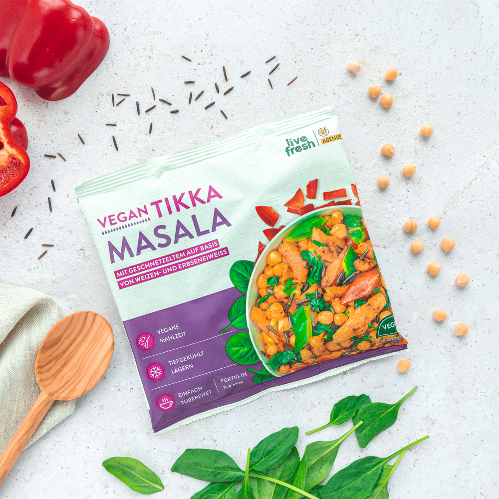 Eine Verpackung des "Vegan Tikka Masala" von LiveFresh, umgeben von frischen Zutaten wie Kichererbsen, Paprika und Spinatblättern. Auf der Verpackung sind Informationen wie "vegane Mahlzeit", "tiefgekühlt lagern" und "einfach zubereitet" zu lesen.