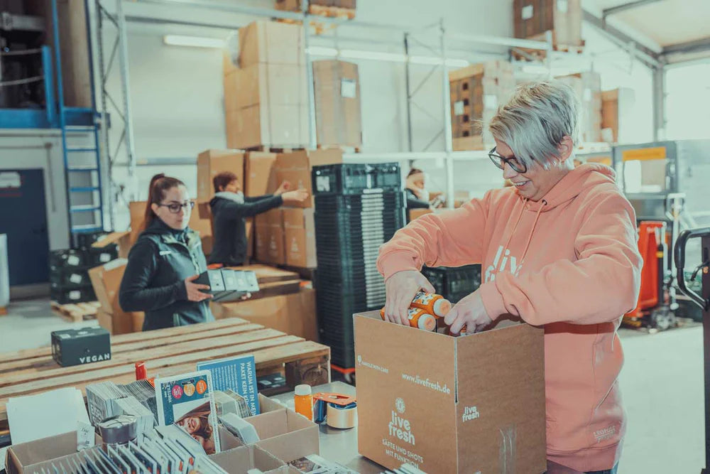 Eine Frau mit kurzen Haaren, die in einem Lager eine Versandbox mit „live fresh“-Produkten befüllt. Sie trägt einen rosa Hoodie mit dem „live fresh“-Logo. Im Hintergrund sind weitere Personen zu sehen, die ebenfalls in der Verpackung und Logistik tätig sind. Kartons und Paletten umgeben den Raum, was auf eine organisierte Produktions- und Versandumgebung hindeutet. Die Szene wirkt geschäftig und professionell, mit einem klaren Fokus auf das sorgfältige Verpacken der Produkte.