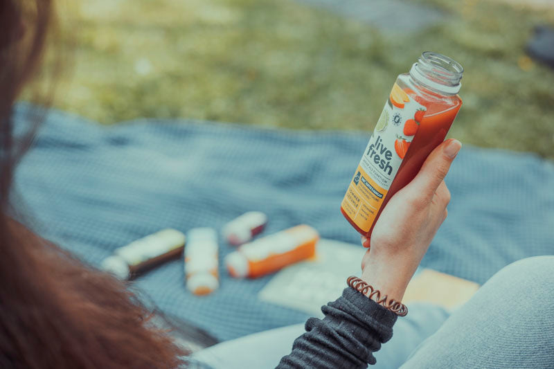 ine Person, die eine Flasche "Live Fresh" Saft in der Hand hält, während sie auf einer Picknickdecke sitzt. Im Hintergrund sind weitere Flaschen und Gegenstände unscharf auf der Decke zu sehen, was eine entspannte Picknick-Atmosphäre vermittelt. 