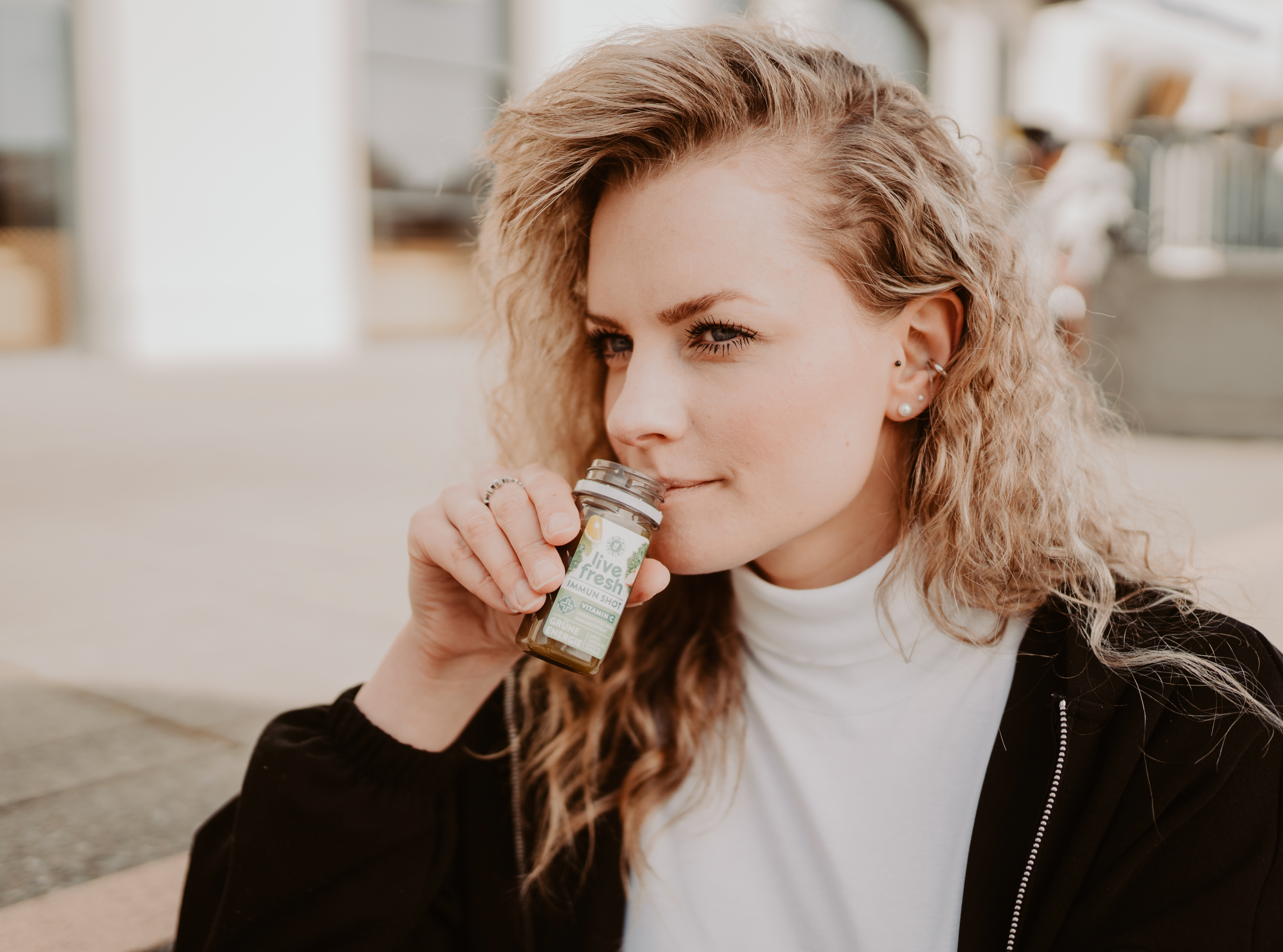 Frau mit lockigem, blondem Haar hält einen Live Fresh Immun Shot an ihre Lippen, bereit, den Shot zu trinken. Sie trägt ein weißes Oberteil und eine schwarze Jacke, während sie draußen sitzt. Im Hintergrund ist eine urbane Umgebung zu erkennen