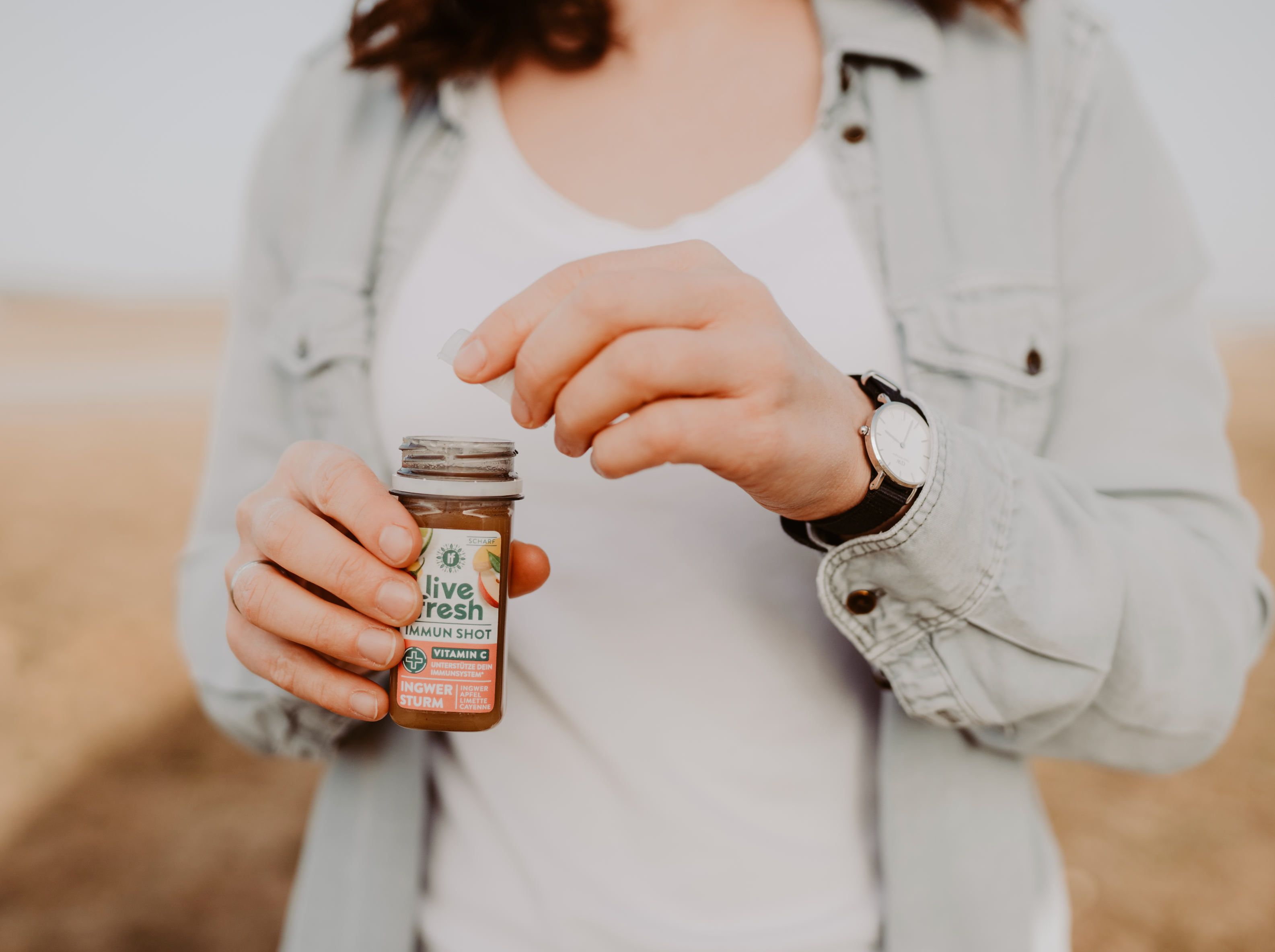 Person öffnet einen Live Fresh Immun Shot mit der Geschmacksrichtung "Ingwer Sturm" und Vitamin C. Sie trägt eine helle Jeansjacke und eine Armbanduhr, während sie den Deckel von der kleinen Shot-Flasche abnimmt. Der Hintergrund deutet auf eine Outdoor-Umgebung hin.