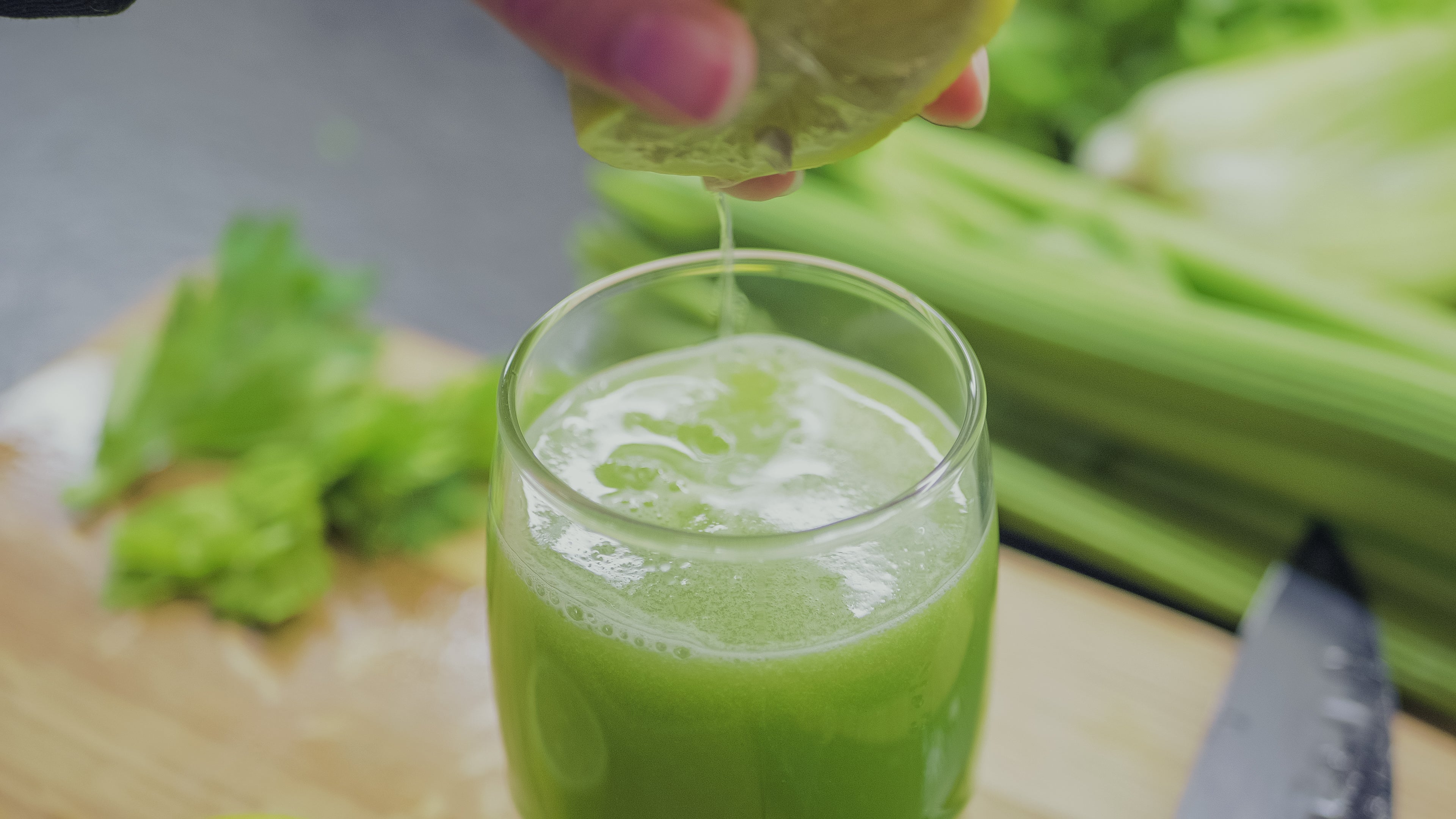 Eine Hand, die eine Limette über einem Glas grünen Selleriesaft auspresst. Im Hintergrund sind frische Selleriestangen und Blätter auf einem Schneidebrett zu sehen. Der Saft im Glas ist schaumig und frisch, was eine natürliche, gesunde und erfrischende Zubereitung von Getränken vermittelt.