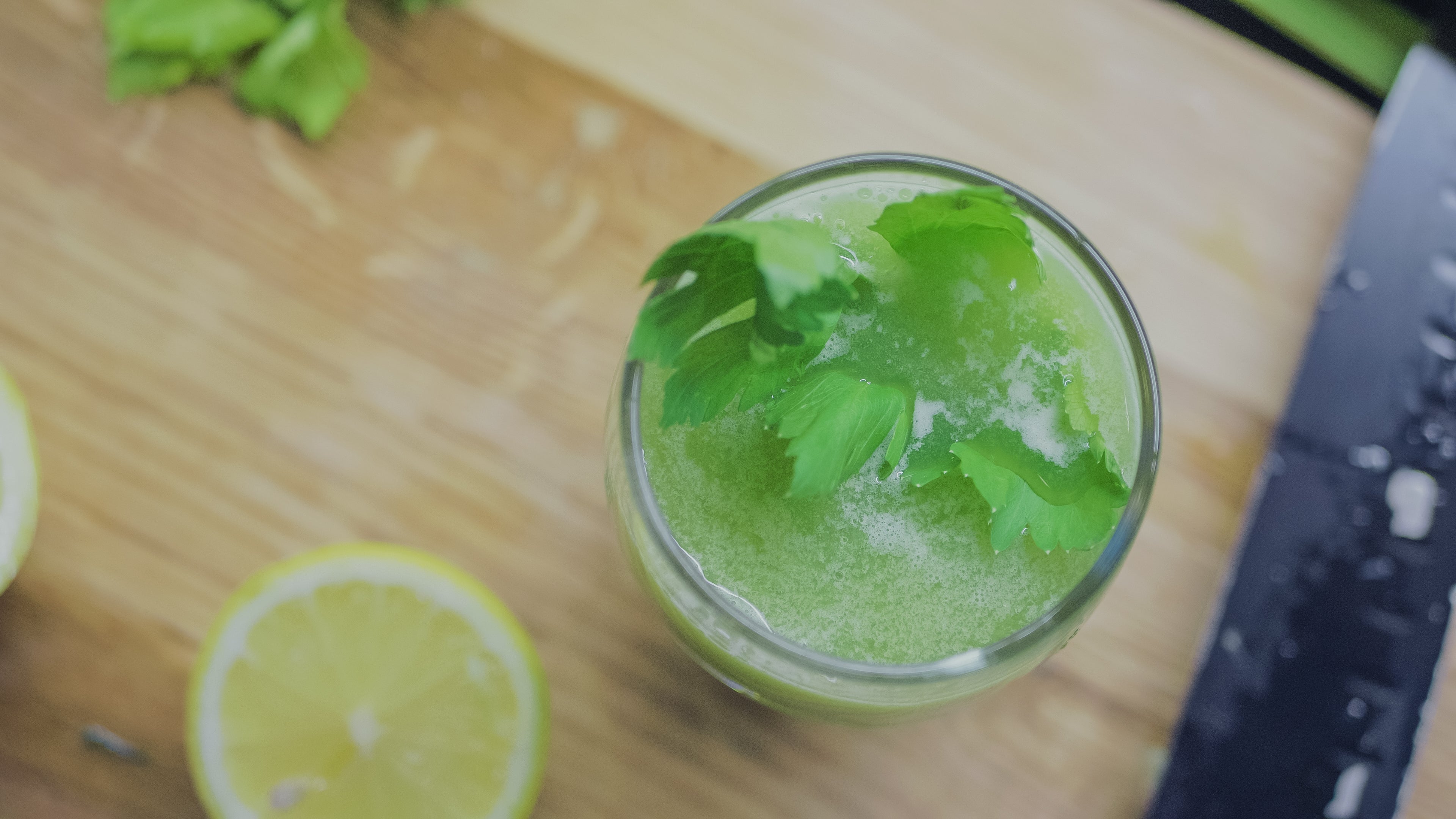 Ein Glas mit grünem Selleriesaft, der mit frischen Sellerieblättern garniert ist. Neben dem Glas liegt eine angeschnittene Zitronenscheibe auf einem Holzuntergrund. Die Szene vermittelt Frische und Gesundheit und deutet auf einen erfrischenden, natürlichen Saft hin.