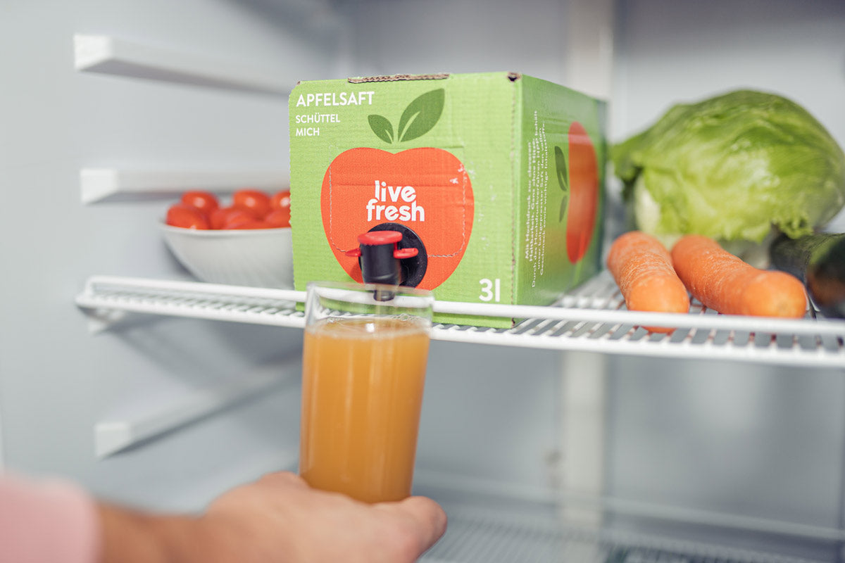 Einen Kühlschrank, in dem eine „Live Fresh“ Apfelsaftbox mit 3 Litern kaltgepresstem Saft steht. Der Zapfhahn der Box wird verwendet, um ein Glas Apfelsaft zu füllen. Neben der Saftbox befinden sich frische Lebensmittel wie Karotten, Salat und eine Schale mit Tomaten. Auf der Box steht „Apfelsaft – Schüttel mich“, was auf die Frische und den kalten Genuss des Safts hinweist.