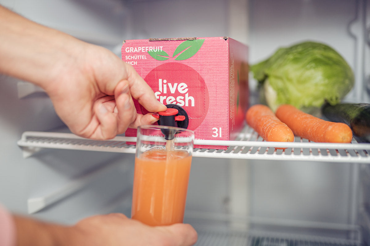 Eine Person, die in einem Kühlschrank ein Glas mit Grapefruitsaft aus einer „Live Fresh“ 3-Liter-Box mit kaltgepresstem Saft zapft. Auf der Saftbox steht „Grapefruit – Schüttel mich“. Neben der Box liegen frische Karotten, Salat und eine Gurke. Der Zapfhahn der Box wird betätigt, um den frischen Grapefruitsaft ins Glas zu füllen.