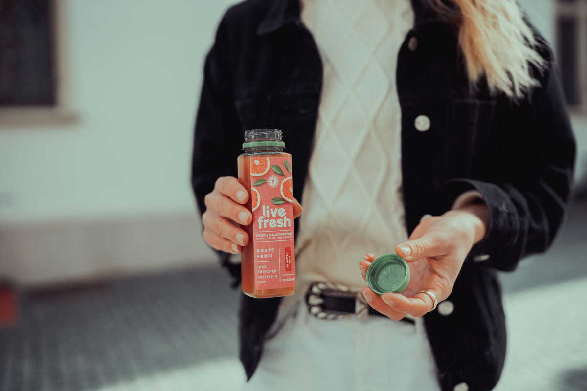 Eine Person, die eine geöffnete Flasche „Live Fresh“ Grapefruit-Saft in der einen Hand und den grünen Deckel der Flasche in der anderen hält. Das Etikett auf der Flasche beschreibt den Saft als „Frisch & Kaltgepresst, Keine Zusätze – Nie Erhitzt“ sowie „100% Frischer Grapefruit Saft“. Die Person trägt eine weiße Strickjacke unter einer schwarzen Jacke und steht in einer hellen Umgebung.