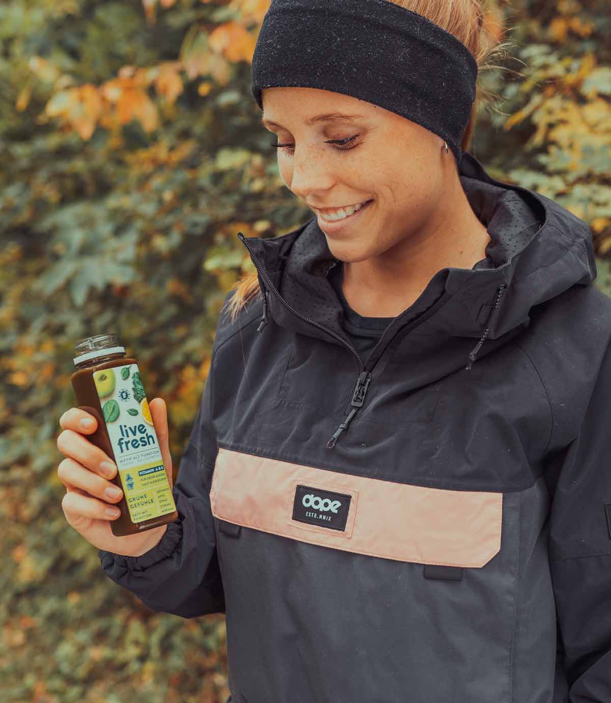 Zeigt eine lächelnde Frau in sportlicher Outdoor-Kleidung, die eine geöffnete Flasche „Live Fresh Grüne Gefühle“ in der Hand hält. Die Szene spielt sich in einer herbstlichen Umgebung ab, was durch die orange-gelben Blätter im Hintergrund betont wird. Die Flasche enthält ein gesundes, kaltgepresstes, veganes Getränk, das Frische und Vitalität symbolisiert.