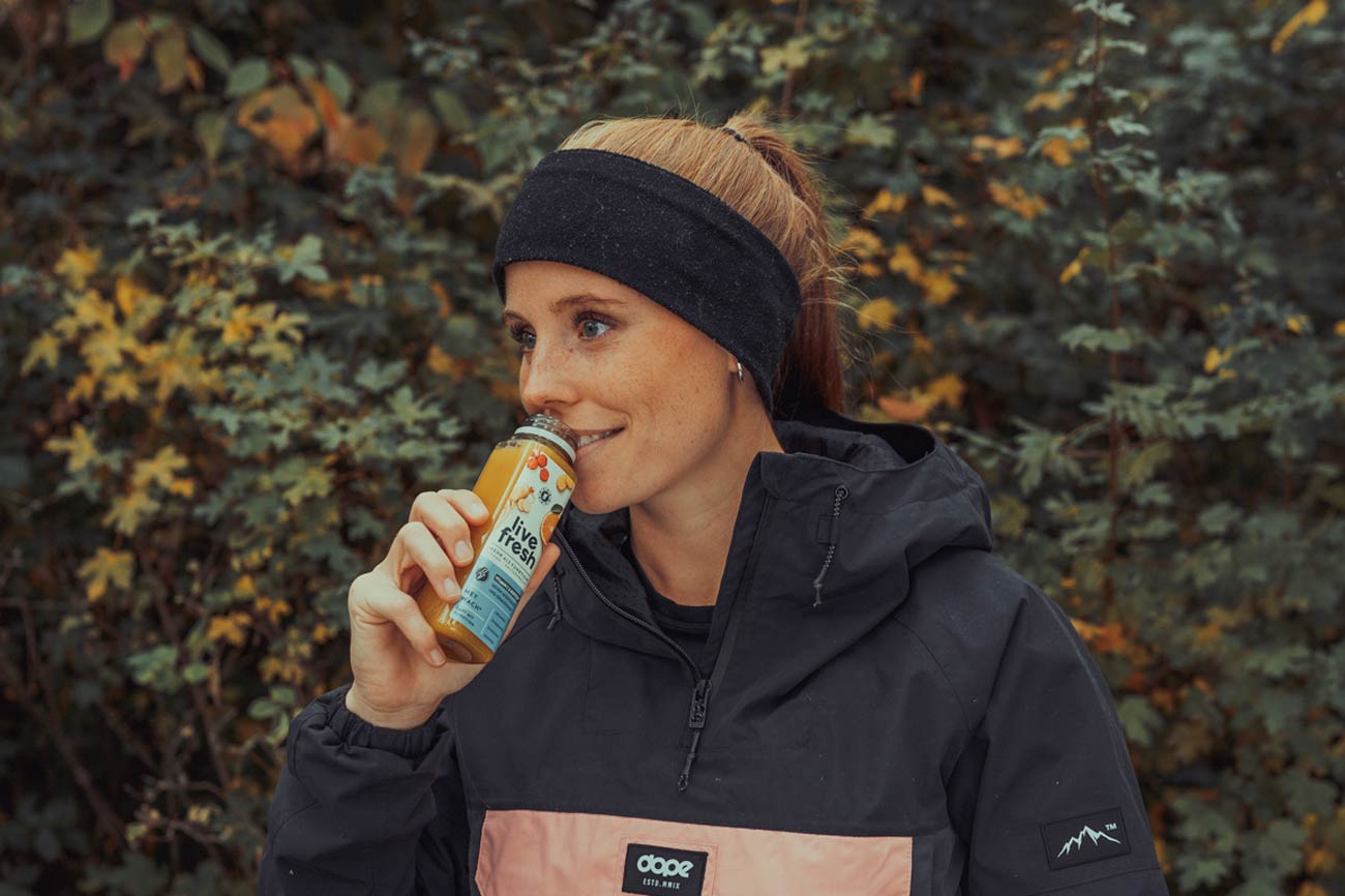 eine Frau, die draußen steht und einen „Live Fresh Hey Wach“ Saft trinkt. Sie trägt eine sportliche Jacke und ein Stirnband, was auf eine Aktivität im Freien hinweist. Im Hintergrund sind grüne Büsche und Bäume zu sehen, die eine herbstliche Stimmung vermitteln. Der „Hey Wach“ Saft in ihrer Hand signalisiert einen gesunden, erfrischenden Energieschub, passend zu einem aktiven Lebensstil.