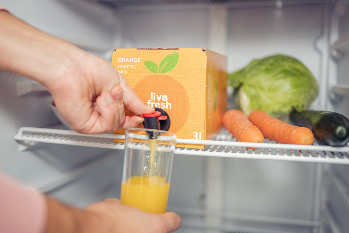 Eine Person, die in einem Kühlschrank ein Glas mit Orangensaft aus einer „Live Fresh“ 3-Liter-Box mit kaltgepresstem Saft zapft. Auf der Saftbox steht „Orange – Schüttel mich“. Neben der Box liegen frische Karotten, Salat und eine Gurke. Der Zapfhahn der Box wird betätigt, um den frischen Orangensaft ins Glas zu füllen.