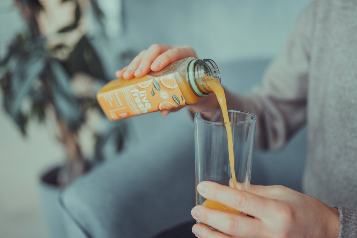 Nahaufnahme einer Person, die einen orangefarbenen Saft aus einer Flasche mit der Aufschrift „Live Fresh“ in ein durchsichtiges Glas gießt. Auf der Flasche sind Orangen abgebildet, und das Etikett bewirbt das Produkt als frischen Saft. Die Szene wirkt entspannt, mit weichem Licht und einer gemütlichen Atmosphäre.